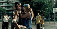 An African American man holding a young blonde woman in her arms. They are standing in a basketball court. The woman is putting her hands into the man’s face.