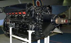 A front right view of a large, black-painted, piston aircraft engine with a prominent propeller shaft. A camouflaged military aircraft is parked behind.