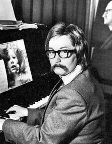На изображении женщина в образе мужчины с усами, в очках, сидящая за пианино