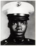 Head of a young black man staring intently at the camera, wearing a dark military jacket and a white peaked cap with a black visor.