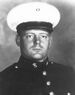 Head and shoulders of a young white man wearing a white peaked cap with a black visor and a dark military jacket with bright buttons down the center.