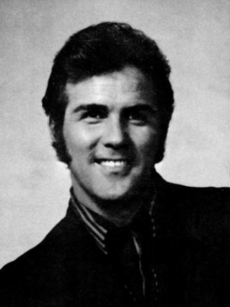 A dark-haired man wearing a shirt and a dark jacket, smiling broadly