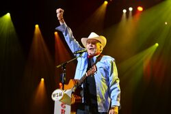 An older man wearing a white cowboy hat and a denim shirt, with a guitar hanging over his shoulder standing at a microphone raising his right fist in the air