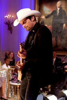 A man wearing a dark jacket and white cowboy hat playing guitar in front of an audience