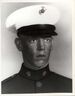 Head of a young white man wearing a white peaked cap with black visor and a dark military jacket with a pin on each side of the upright collar.