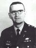 Head and shoulders of a middle-aged man with closely cropped hair and horn-rimmed glasses. He is wearing a dark military jacket with a parachute badge on the left breast and a Christian cross under a "U.S." pin on each lapel.