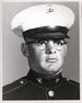 Head of a heavyset young white man with wire-frame glasses wearing a white peaked cap with black visor and a dark military jacket.