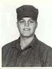 Head and shoulders of a smiling young white man wearing a cloth army cap and a plain buttoned-down shirt of the same color.