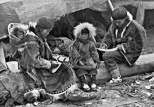 An Inuit family is sitting on a log outside their tent. The parents, wearing warm clothing made of animal skins, are engaged in domestic tasks. Between them sits a toddler, also in skin clothes, staring at the camera. On the mother’s back is a baby in a papoose.