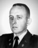 Head of a solemn-faced young man with short hair wearing a dark military jacket with pins on the lapels over a shirt and tie.