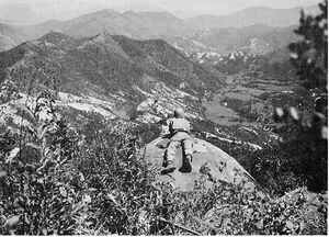 A man lays on a cliff overlooking a large hill complex