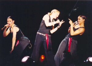 Picture of three woman. The one in the middle has her blonde hair tied in a ponytail. She holds a microphone on her left arm and is leaning towards her right and pointing towards an African American female who’s pointing back at her. On the left there’s a caucasian female holding a microphone with her left hand.
