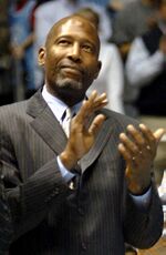 A man, wearing a gray suit, a shirt and a tie, is clapping his hands while looking up.