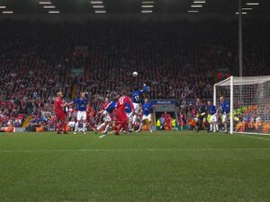 People in blue and red shirts on a field with a ball in the air. In the background is a stand that contains a lot of people.