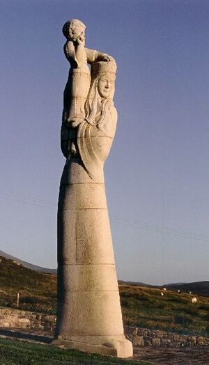 Большая каменная статуя высокой и худой длинноволосой женщины, возвышающаяся над полем. Женщина одета в платье и головной убор, на плече у неё стоит ребёнок, которого она поддерживает