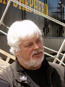 A heavyset man with white hair and a white beard, standing in front of a boat with the words «Shepherd» written on it in yellow paint