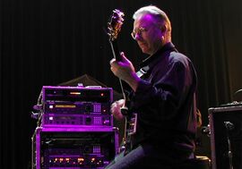 An older man from the side, holding a guitar with an amp to his left under a purple spotlight