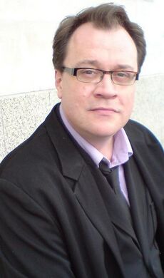Мужчина в очках, одетый в чёрный пиджак, жилетку, розовую рубашку, галстук и джинсы, сидит на фоне стены мраморного цвета