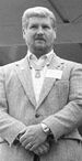 A white man with thick hair and a closely trimmed beard standing with his hands crossed in front of him. He is wearing a suit coat with a star-shaped medal hanging from a ribbon around his neck.