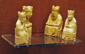 Четыре золотые антропоморфные шахматные фигуры, стоящие на стеклянной полке на фоне красной стены. Все фигуры сидят на стульях, на головах у них короны. Две фигуры большего размера — мужские, с бородами; остальные женские