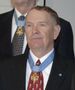 Head and shoulders of an older man with thinning hair wearing a dark suit coat, white shirt, and red tie. A star-shaped medal hangs from a light blue ribbon around his neck and on his lapel is a small round blue pin.